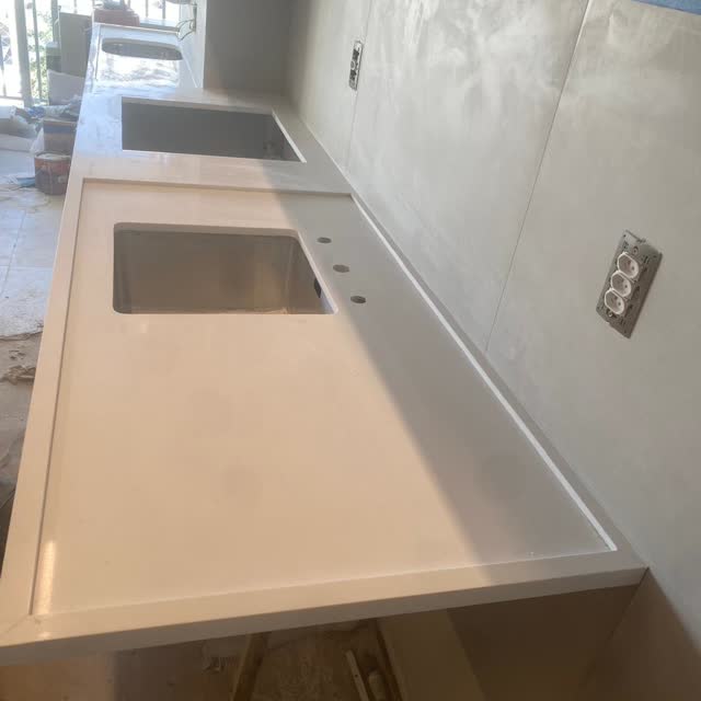 Bancada de cozinha em peça única: Bancada longa em pedra clara com duas cubas esculpidas e furação para torneira, instalada em apartamento em obra.