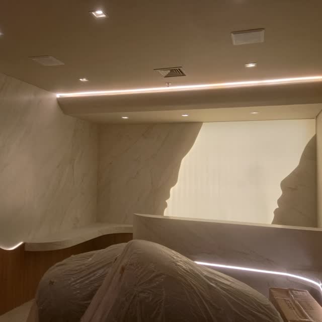 Suíte com painel curvo: Painel de mármore claro com recorte curvo e iluminação embutida, contornando a cabeceira da suíte.