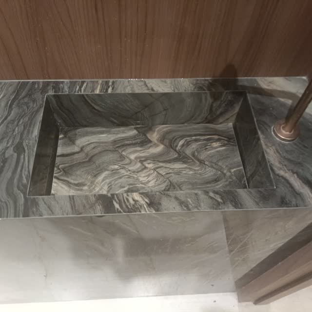 Bancada cinza com cuba esculpida: Bancada em pedra cinza de veio aberto, cuba escavada na peça e metais em ouro velho.