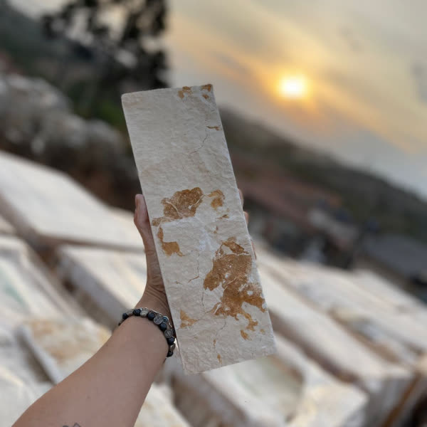 Mão segurando um filete de pedra natural clara contra o sol do fim da tarde, com chapas empilhadas ao fundo.